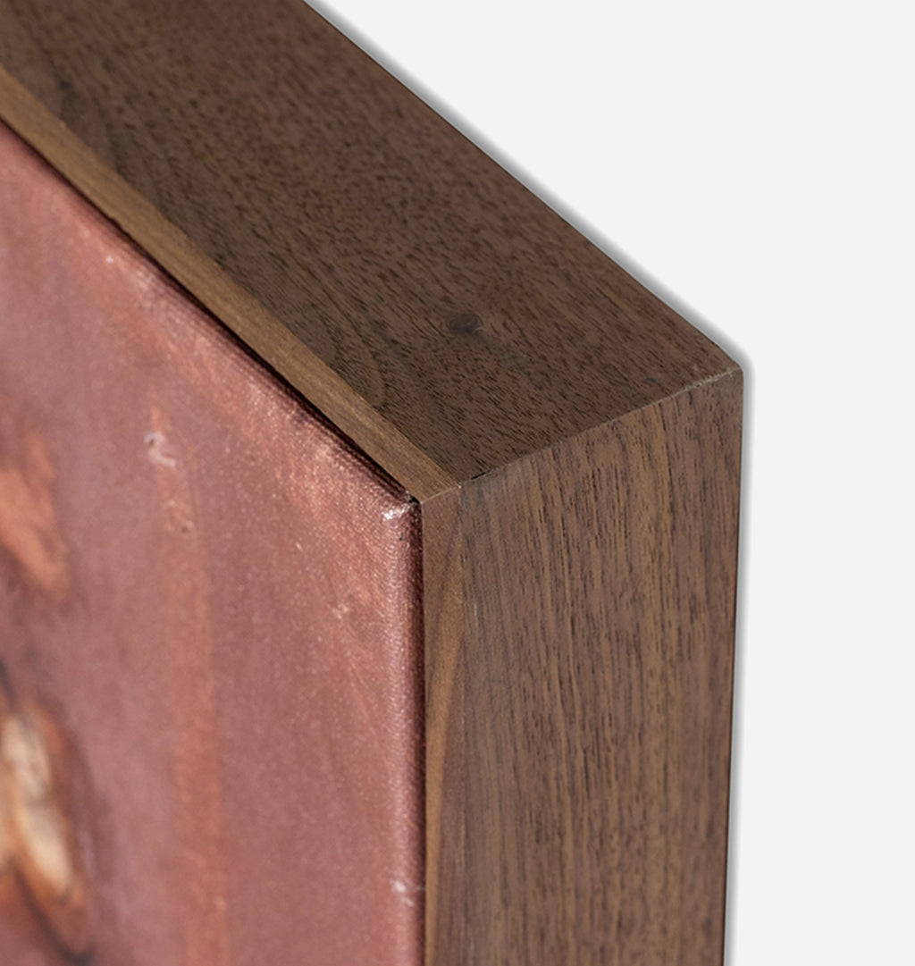 Close-up detail of a framed artwork corner, showing a dark brown wood frame with a textured reddish-brown canvas or print surface against a light gray background.