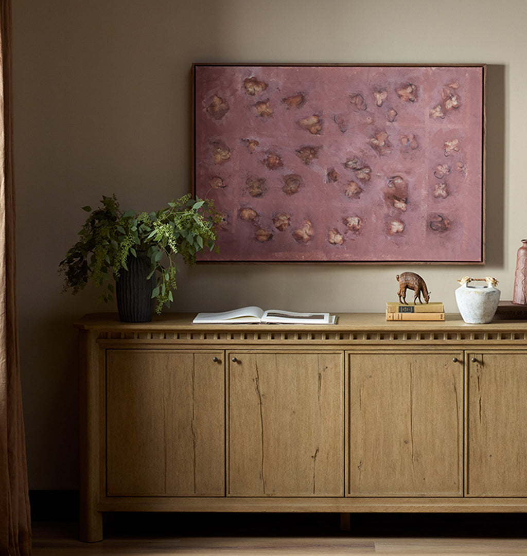 A large framed abstract painting with a dusty rose and brown floral pattern hangs on a beige wall above a light wood credenza in a styled room setting.