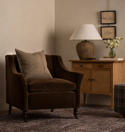 A dark brown velvet armchair with a textured beige throw pillow, positioned next to a wooden sideboard with a lamp and framed art in a warm, neutral-toned room.