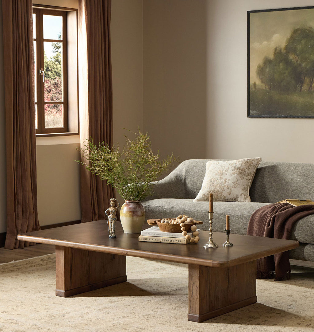 A modern wooden coffee table in a warm brown finish, styled in a cozy living room setting with a gray sofa, brown curtains, and a beige rug.