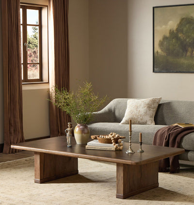 A modern wooden coffee table in a warm brown finish, styled in a cozy living room setting with a gray sofa, brown curtains, and a beige rug.
