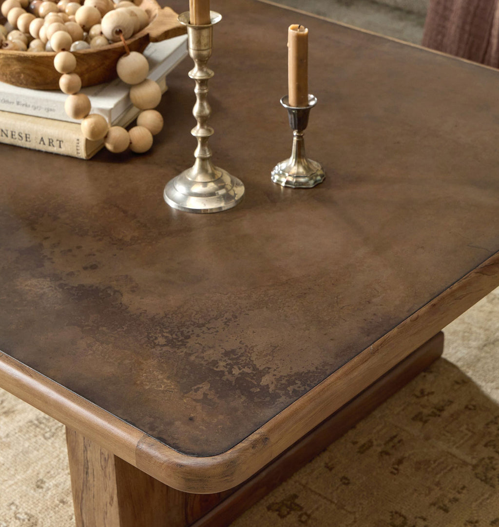 A close-up of a rustic wooden coffee table with a distressed brown top, styled with silver candlesticks, candles, books, and a bowl of wooden beads in a home interior setting.