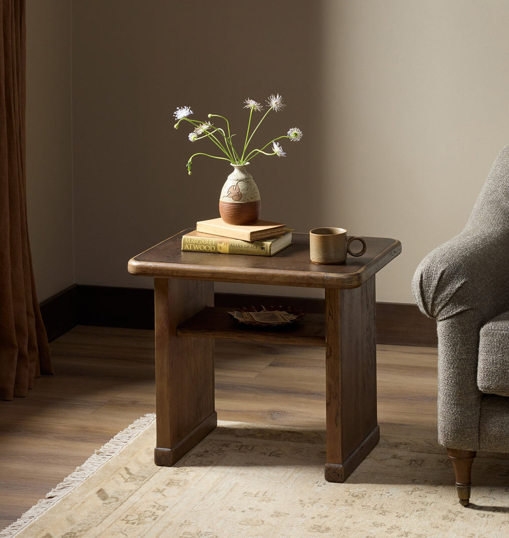 A rustic dark brown wooden end table styled in a cozy living room setting, holding a ceramic vase with wildflowers, a stack of books, and a coffee mug, next to a gray armchair.