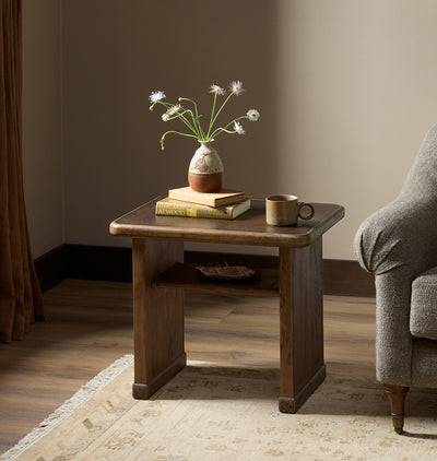 A rustic dark brown wooden end table styled in a cozy living room setting, holding a ceramic vase with wildflowers, a stack of books, and a coffee mug, next to a gray armchair.