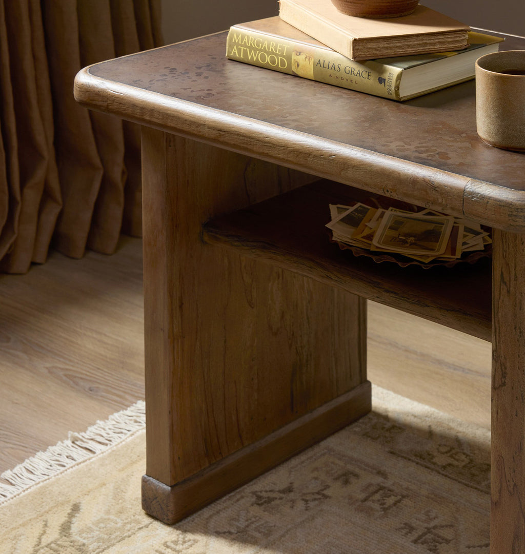 A rustic wooden side table with a thick apron leg and open shelf, styled in a warm room setting with books, a ceramic mug, and curtains in the background.