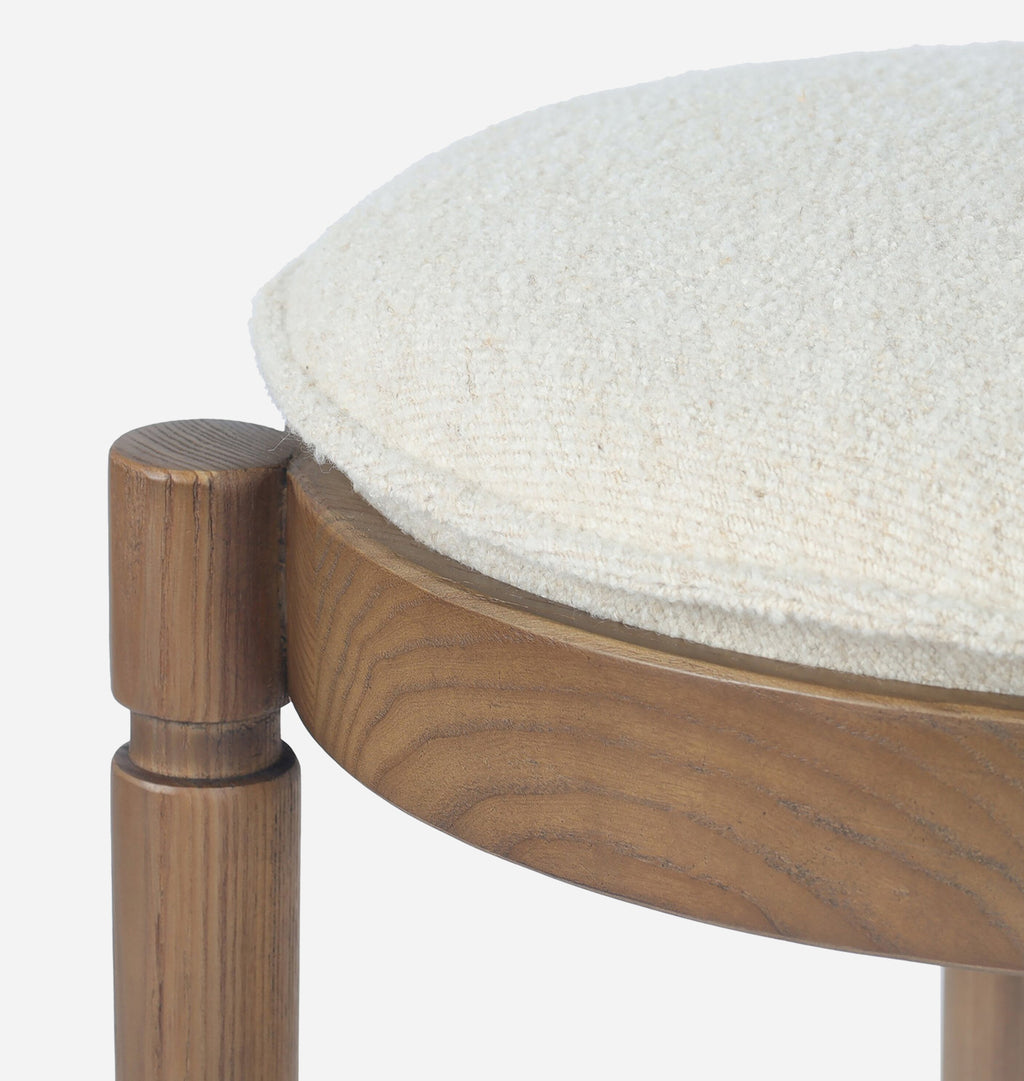 Close-up detail of a wooden stool with a round off-white textured upholstered seat cushion and medium brown wood frame, set against a plain white background.