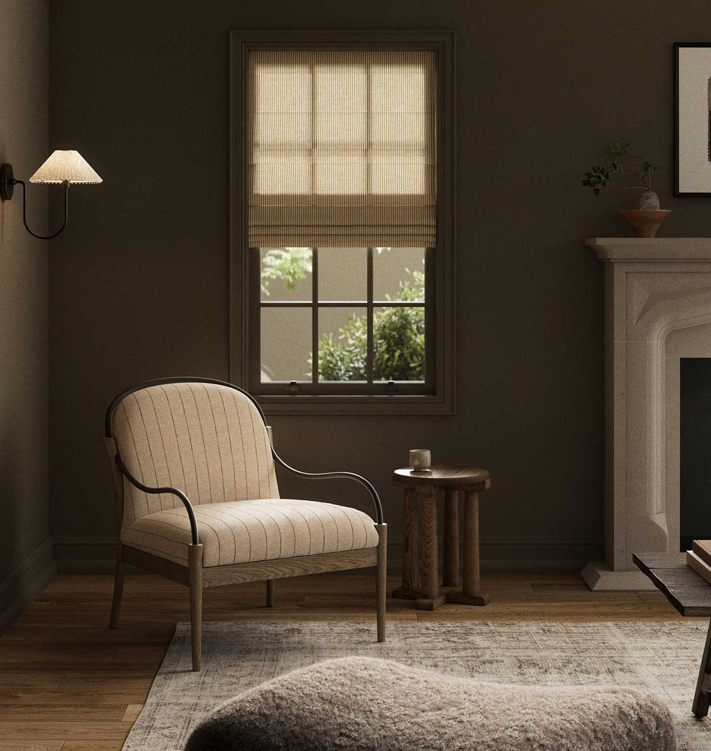 A styled interior room scene featuring a cream ribbed Roman blind on a window, a matching cream upholstered armchair with dark metal arms, a small wooden side table, a fireplace, and a rug in a dark warm-toned room.