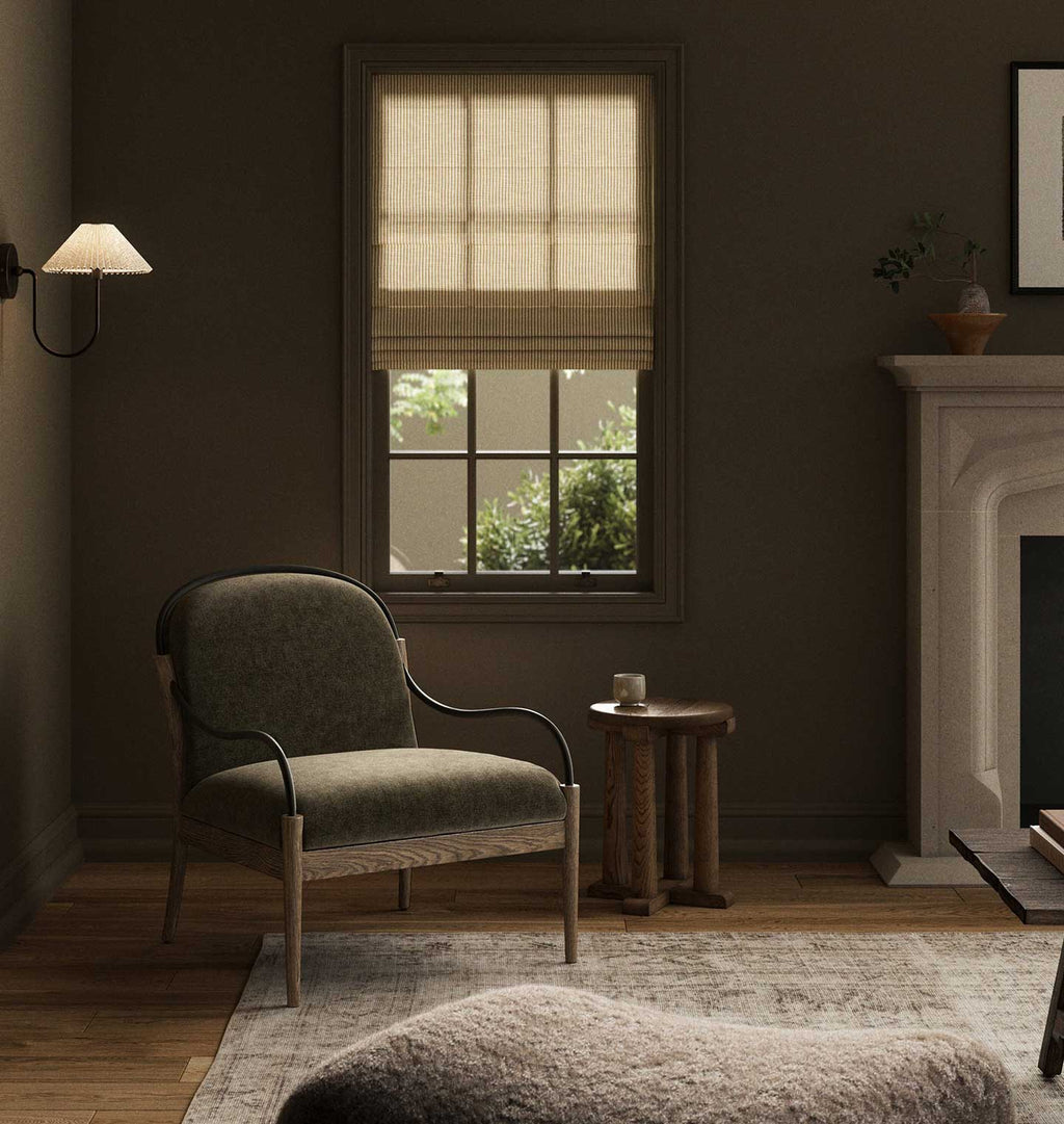 A styled interior room with a window featuring a beige roller blind, a green upholstered armchair, a small wooden side table, a rug, and a fireplace in the background.