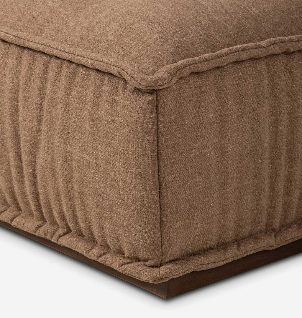 Close-up of a brown upholstered ottoman or bench corner, showing the fabric texture, pleated side panel, and dark wood base on a white background.