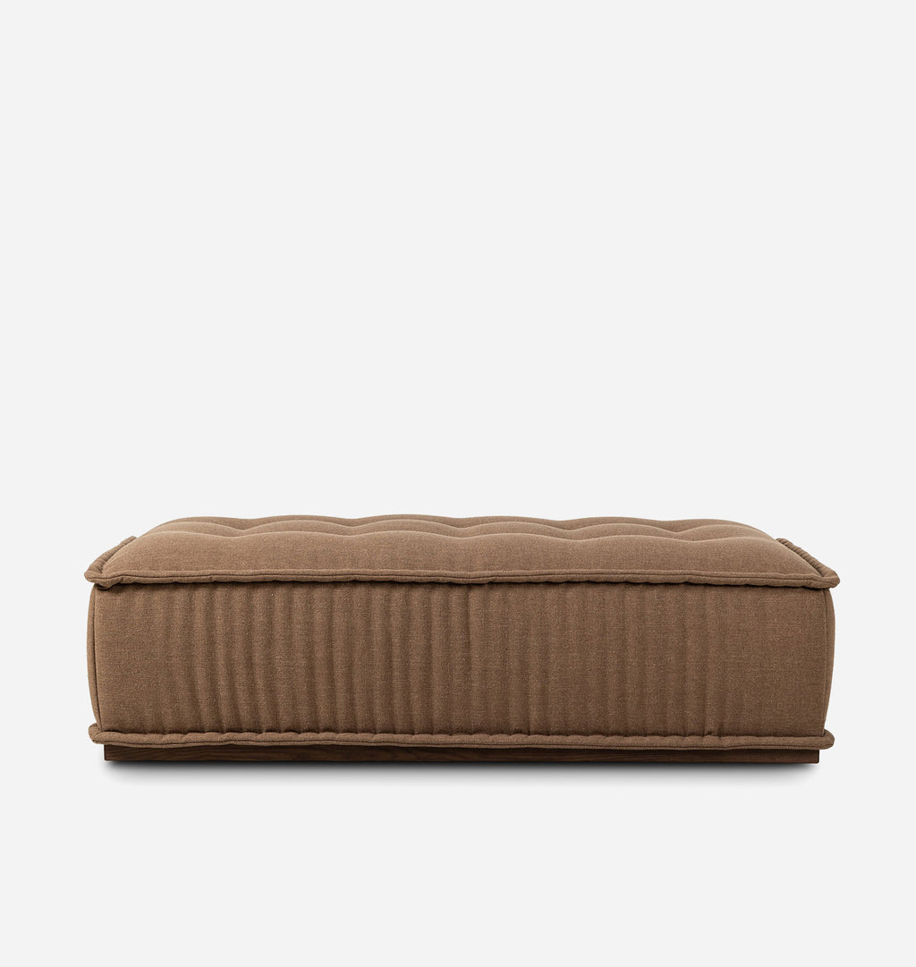 A rectangular upholstered ottoman or bench in a warm taupe-brown fabric, featuring a tufted top and vertical channel ribbing on the sides, shown against a plain light gray background.