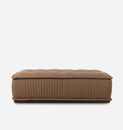 A rectangular upholstered ottoman or bench in a warm taupe-brown fabric, featuring a tufted top and vertical channel ribbing on the sides, shown against a plain light gray background.