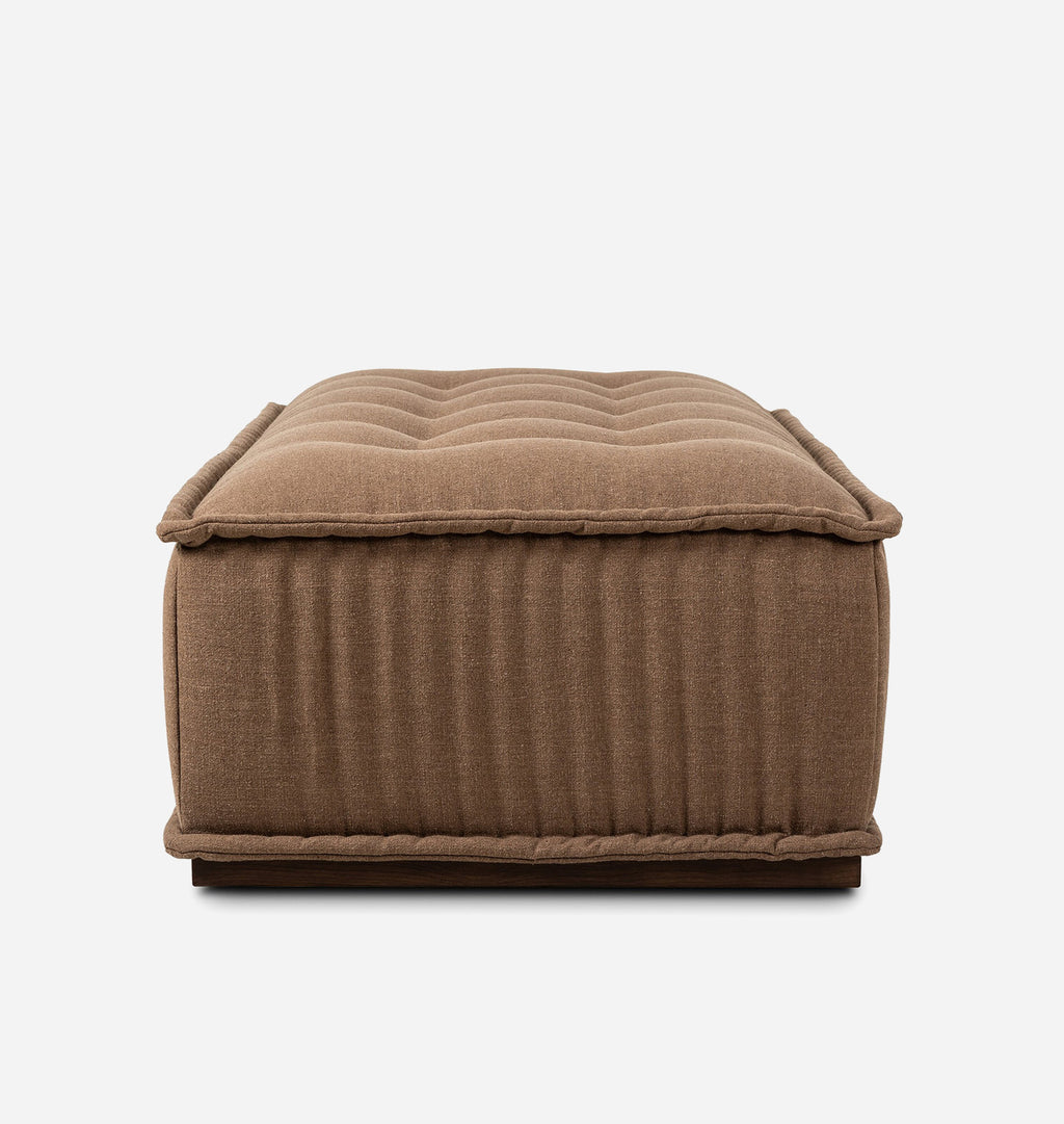A rectangular upholstered ottoman in a warm brown fabric, featuring a tufted top cushion, vertical channel tufting on the sides, and a dark wooden base, shown against a plain white background.