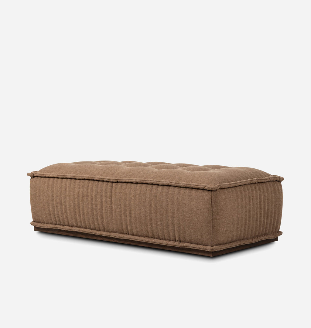 A rectangular upholstered ottoman or bench in a warm taupe-brown fabric, featuring a tufted top and vertical channel ribbing on the sides, shown against a plain light gray background.
