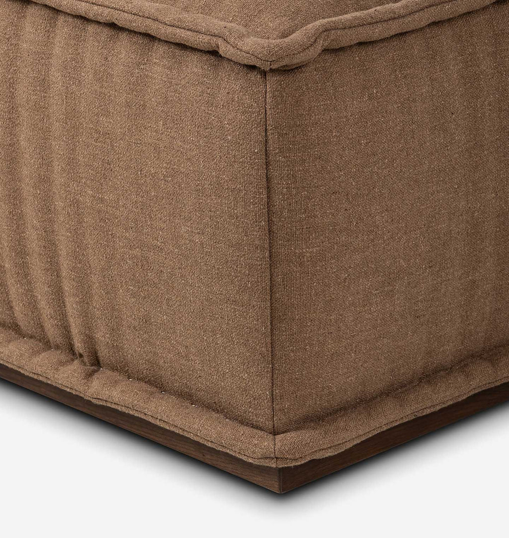 Close-up detail of a brown upholstered ottoman or bench corner, showing the textured fabric, stitched seams, and a dark wood base on a white background.