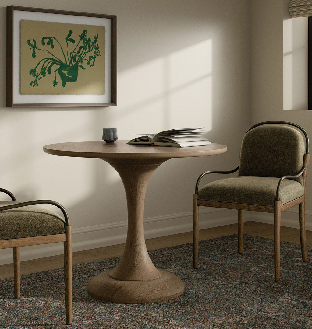 A round wooden dining table with a sculptural pedestal base, styled in a warm, sunlit room with two upholstered wooden chairs, a framed botanical print, and an open book on top.