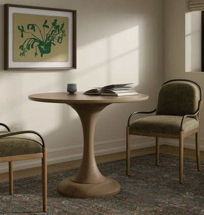 A round wooden dining table with a sculptural pedestal base, styled in a warm, sunlit room with two upholstered wooden chairs, a framed botanical print, and an open book on top.