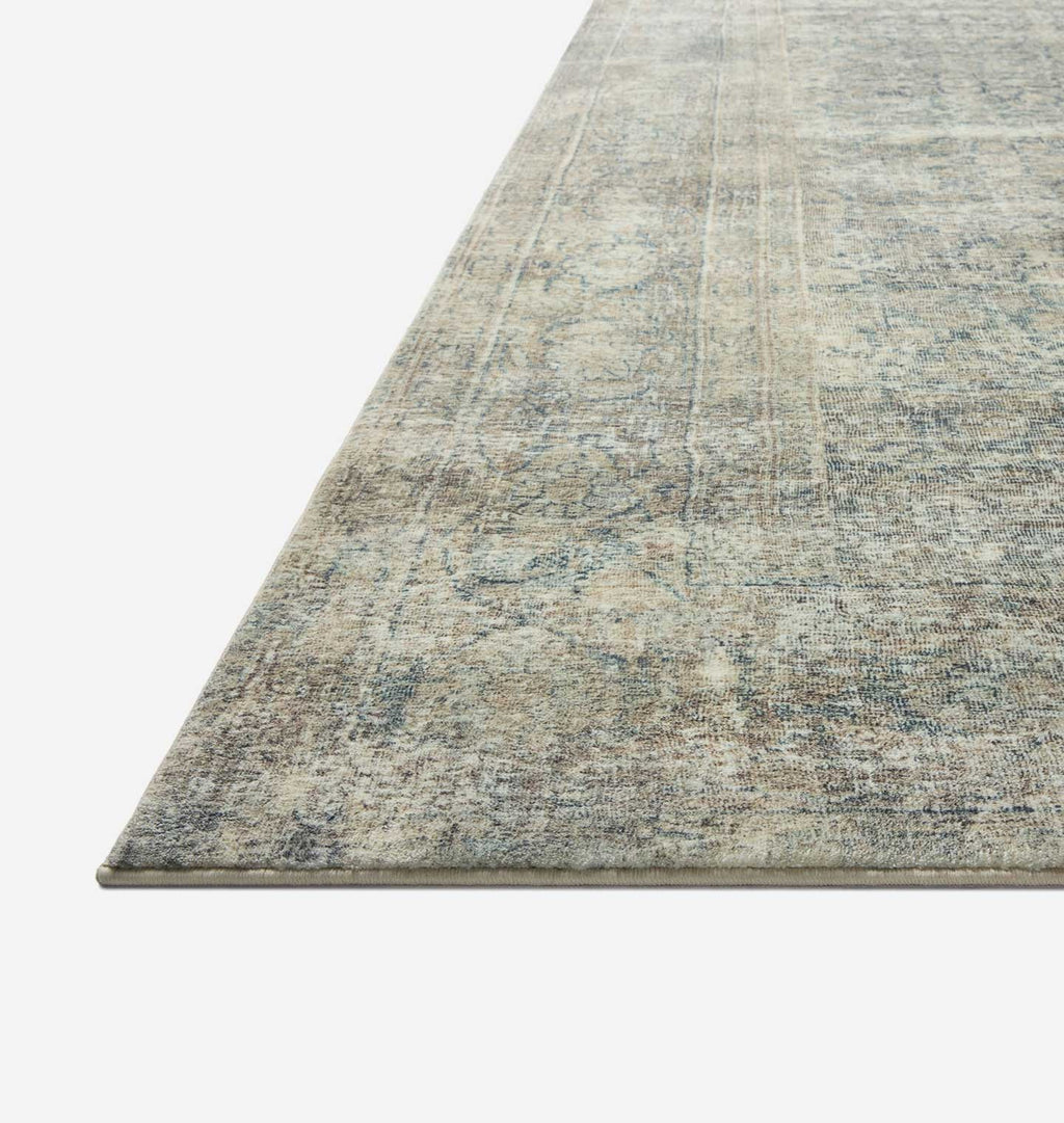 A close-up view of a rectangular area rug with a distressed, vintage-style pattern in muted gray, beige, and blue tones, shown against a plain white background.