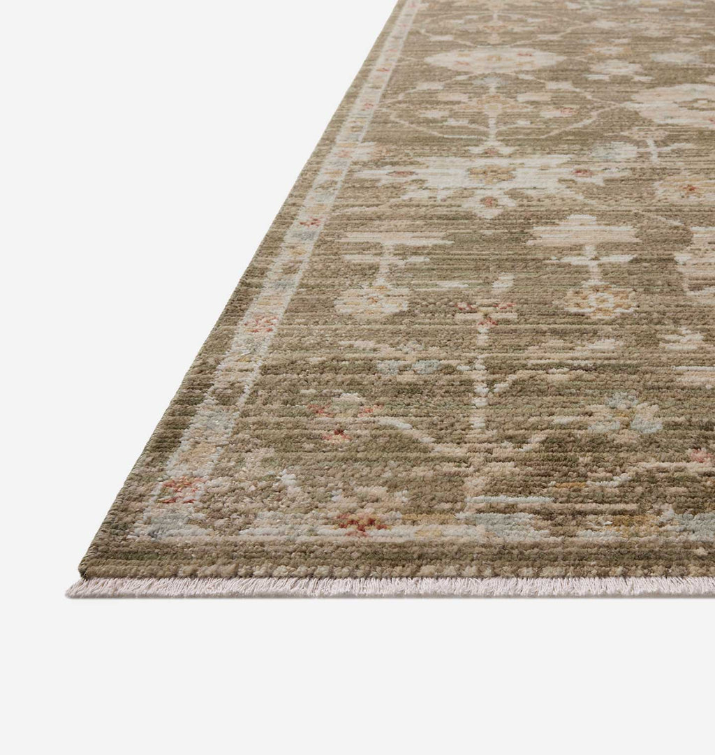 A close-up view of an area rug corner showing its textured fringe and a distressed traditional pattern in muted beige, taupe, and soft pink tones.