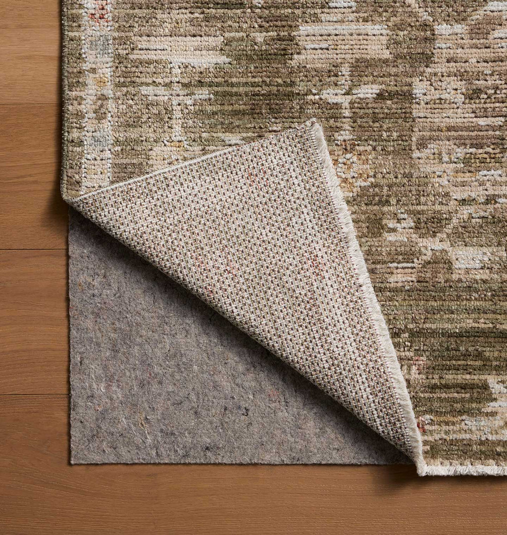A close-up of a textured area rug with a distressed abstract pattern in beige, brown, and muted green tones, shown with one corner folded over to reveal a gray backing on a wooden floor.