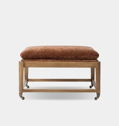 A low wooden bench or ottoman with a thick rectangular cushion upholstered in brown textured fabric, set on casters against a light gray background.