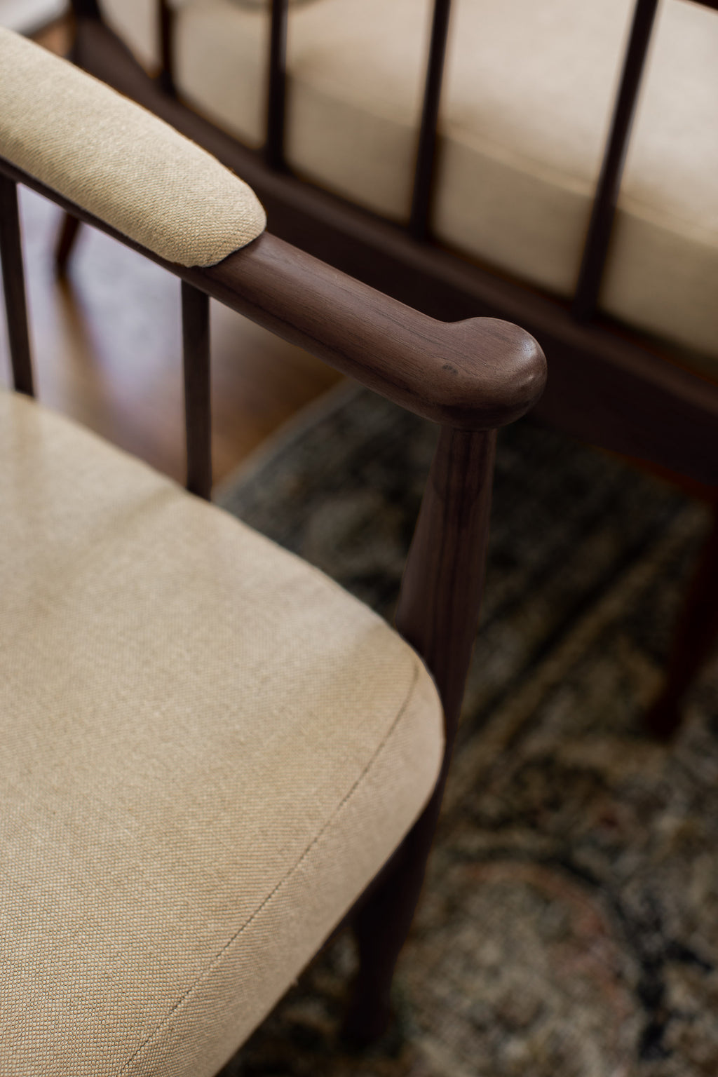A close-up of a mid-century modern armchair with a dark brown wooden frame and beige textured fabric upholstery, set on a patterned rug.