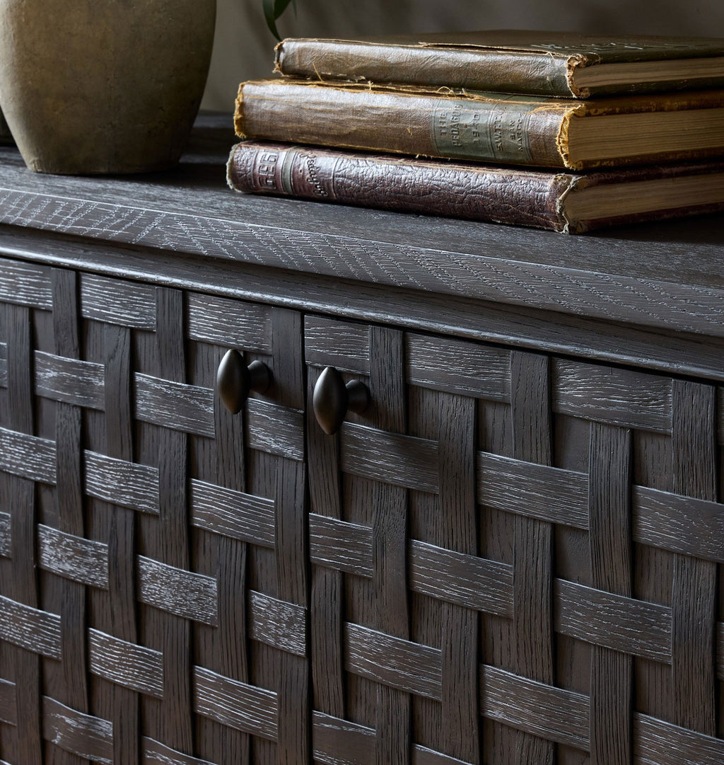 A close-up of a dark wood cabinet with a woven basket-style front, two round dark metal handles, a dark wood top, and a stack of vintage books on top.