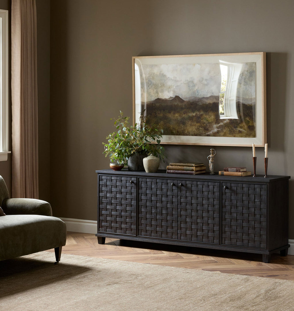 A dark charcoal woven wood credenza in a styled living room setting, decorated with a potted plant, books, and a candle, beneath a large framed landscape print.