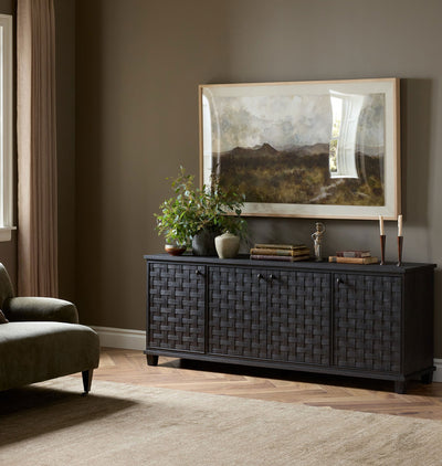 A dark charcoal woven wood credenza in a styled living room setting, decorated with a potted plant, books, and a candle, beneath a large framed landscape print.