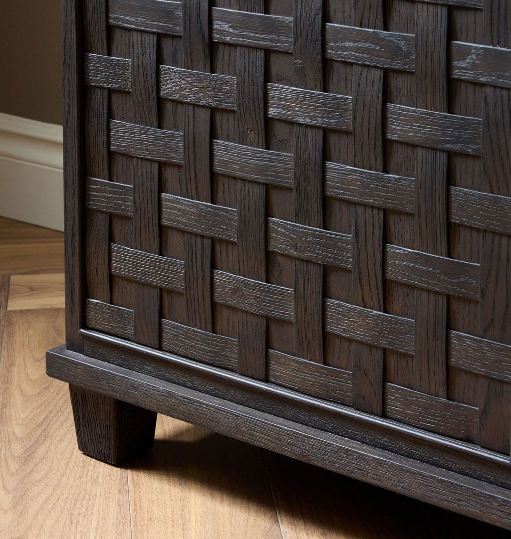 Close-up of a dark brown wooden cabinet with a woven basket-style front panel and block legs, shown on a light wood floor.