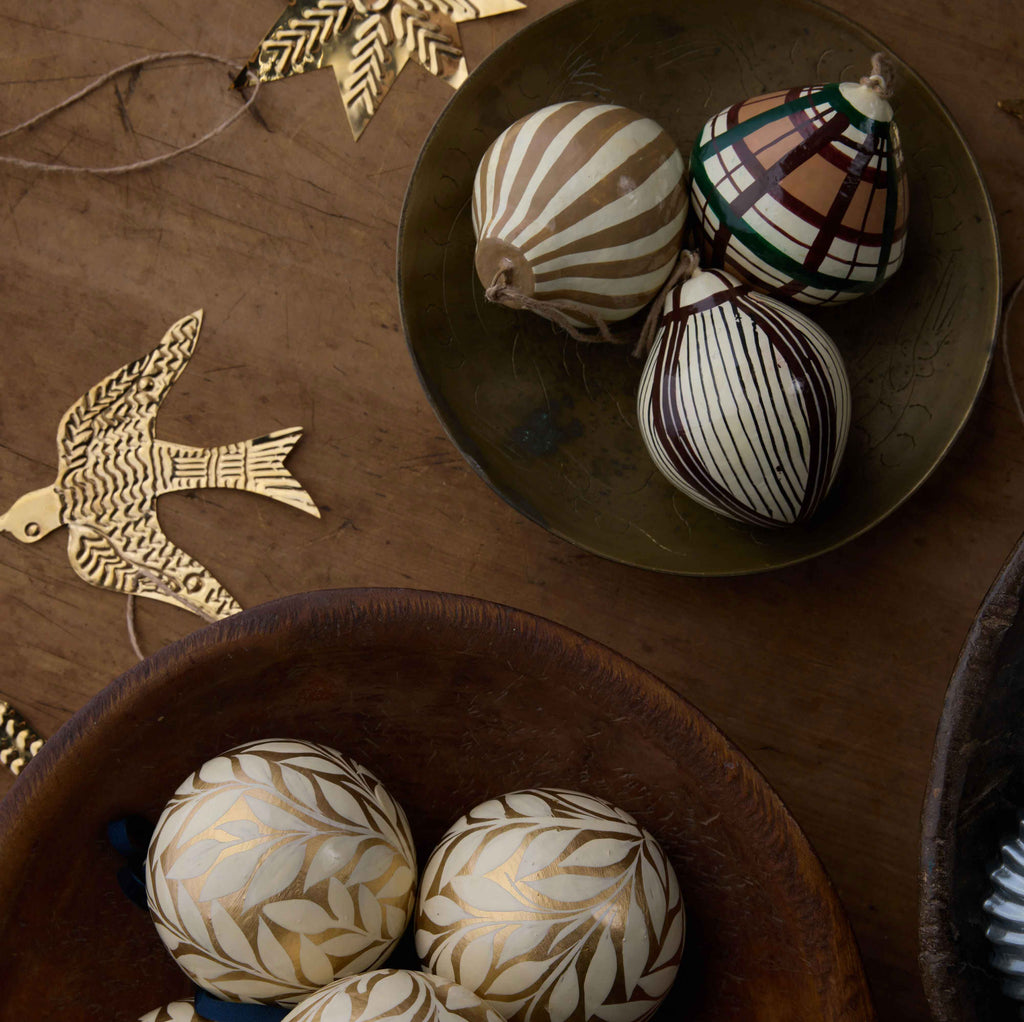 Decorative hand-carved wooden ornaments with intricate leaf and geometric patterns, displayed in rustic bowls on a wooden surface alongside gold-toned bird cutouts.