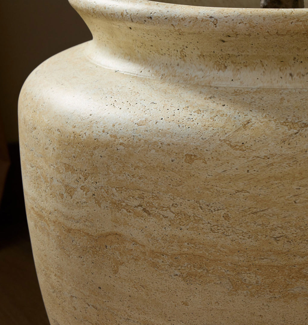 Close-up of a large textured stone planter or vase in a warm beige travertine-like finish, showing the mottled natural stone pattern and curved shoulder detail.