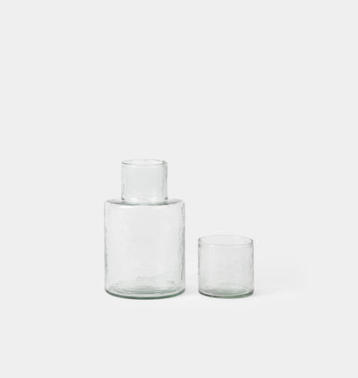 Two clear textured glass vessels, one larger bottle with a short neck and one smaller tumbler, on a light gray background.