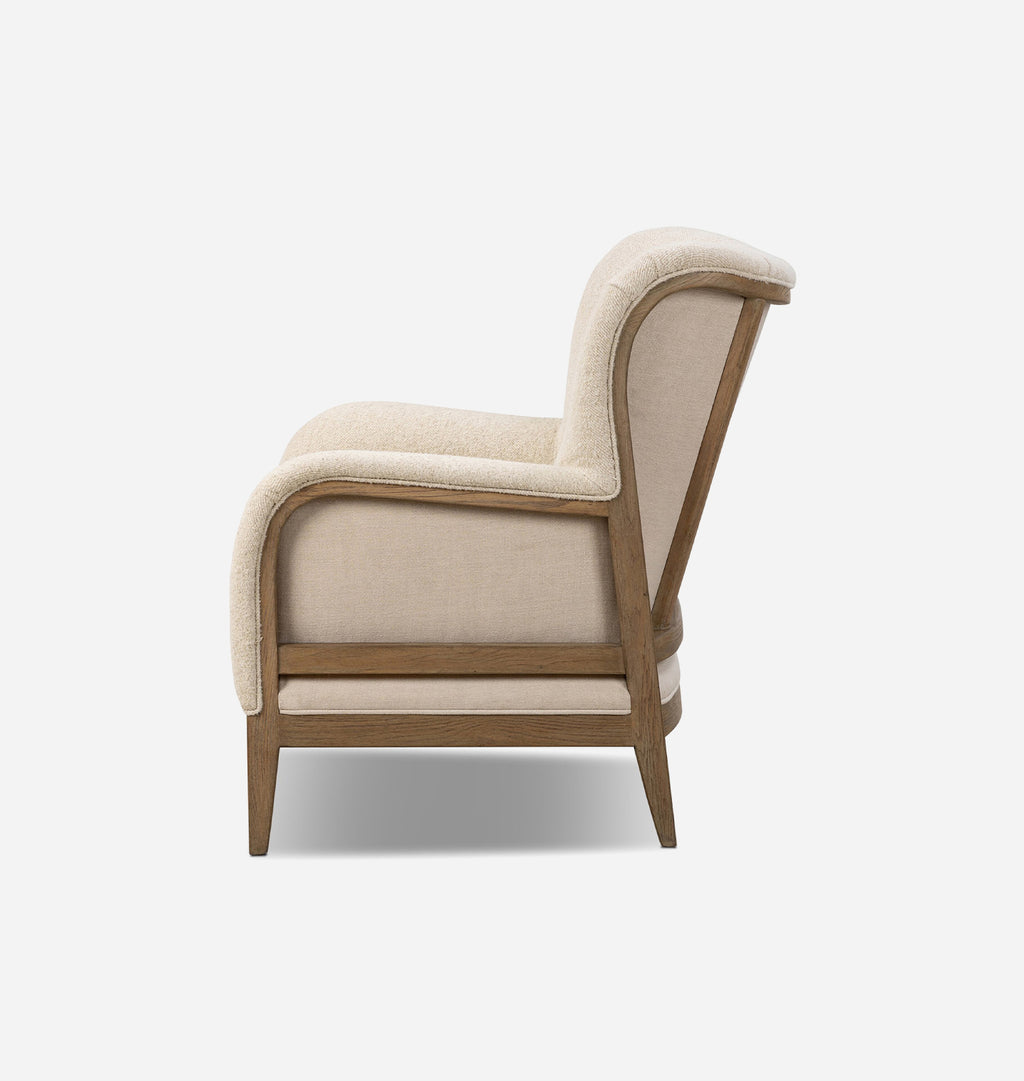 A modern accent armchair with a curved silhouette, upholstered in a textured cream fabric and featuring a medium brown wooden frame and legs, shown from a side profile on a light gray background.