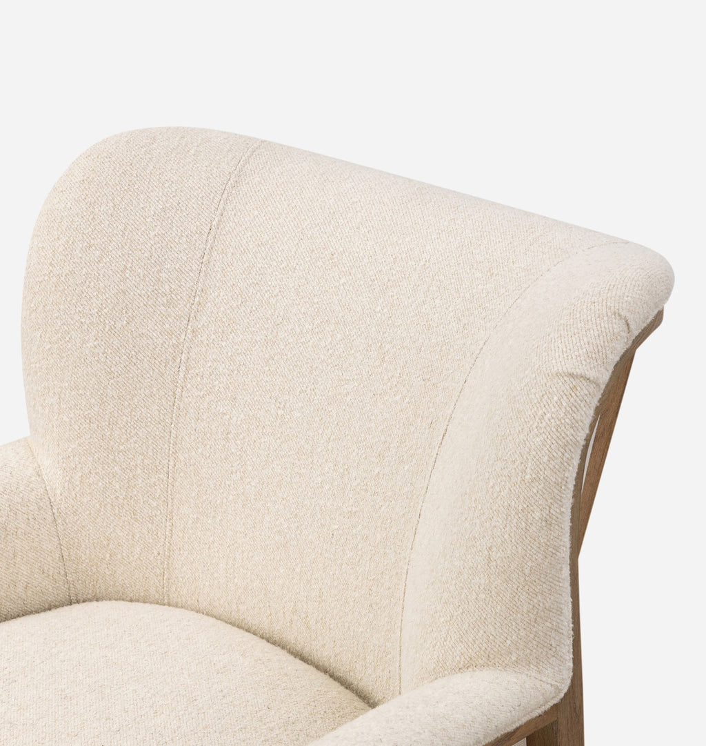 Close-up of a cream-colored upholstered armchair showing the textured fabric, curved backrest, and a bit of the wooden frame against a plain white background.