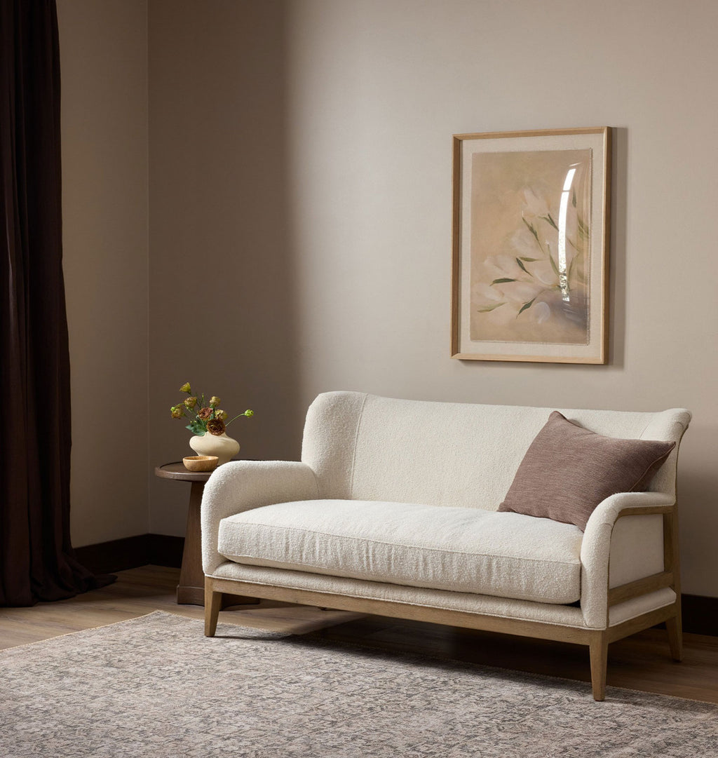 A cream-colored upholstered loveseat with a light wood frame and a brown accent pillow, styled in a minimalist living room setting with a framed print on the wall and a side table.