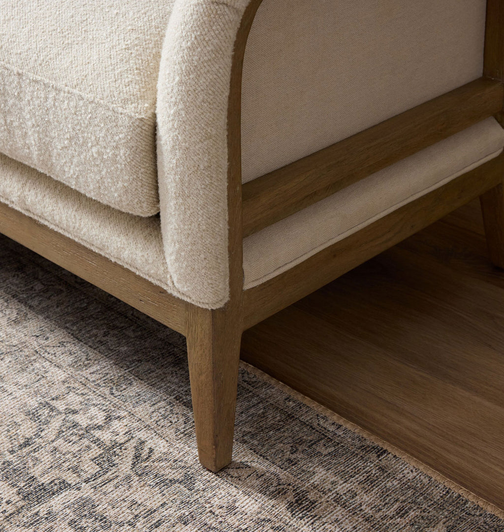 Close-up of a mid-century modern armchair corner with textured cream upholstery and a natural wood frame, shown on a patterned rug over a wood floor.