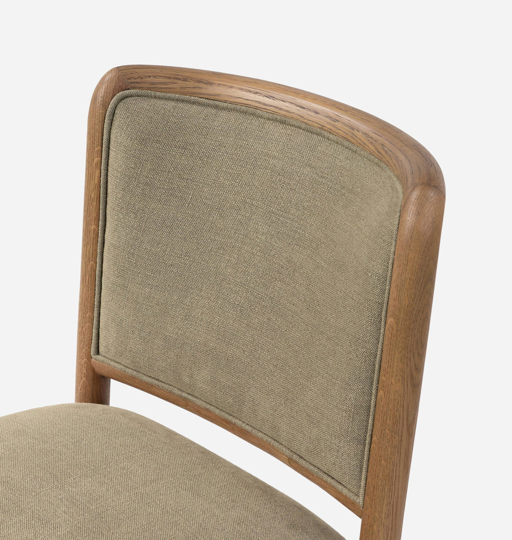 Close-up of a wooden dining chair with a curved upholstered backrest and seat in a warm taupe fabric, set against a plain white background.
