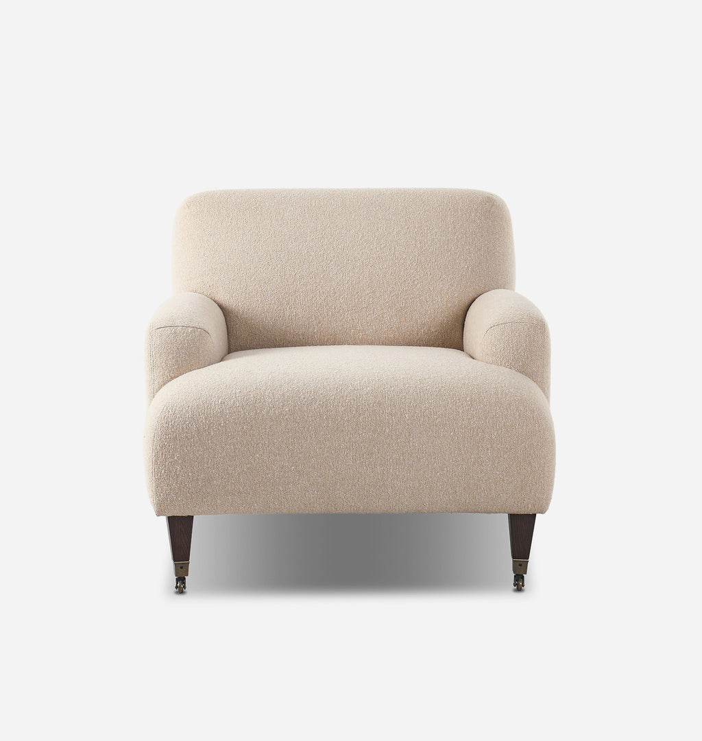 A modern armchair upholstered in a textured beige bouclé fabric, featuring dark wood legs with brass casters, shown against a plain white background.