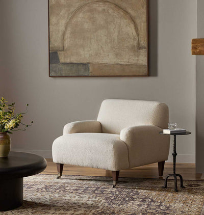 A cream bouclé armchair with dark wooden legs in a styled living room setting, featuring a patterned rug, a small black side table, and an abstract painting on the wall.