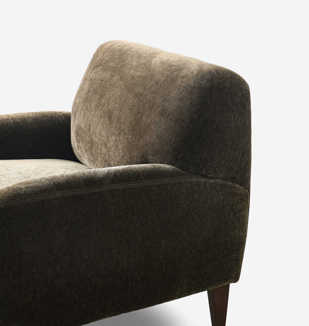 Close-up of the corner of a dark olive upholstered armchair with a textured fabric, showing the backrest, arm, and a portion of the wooden leg against a white background.
