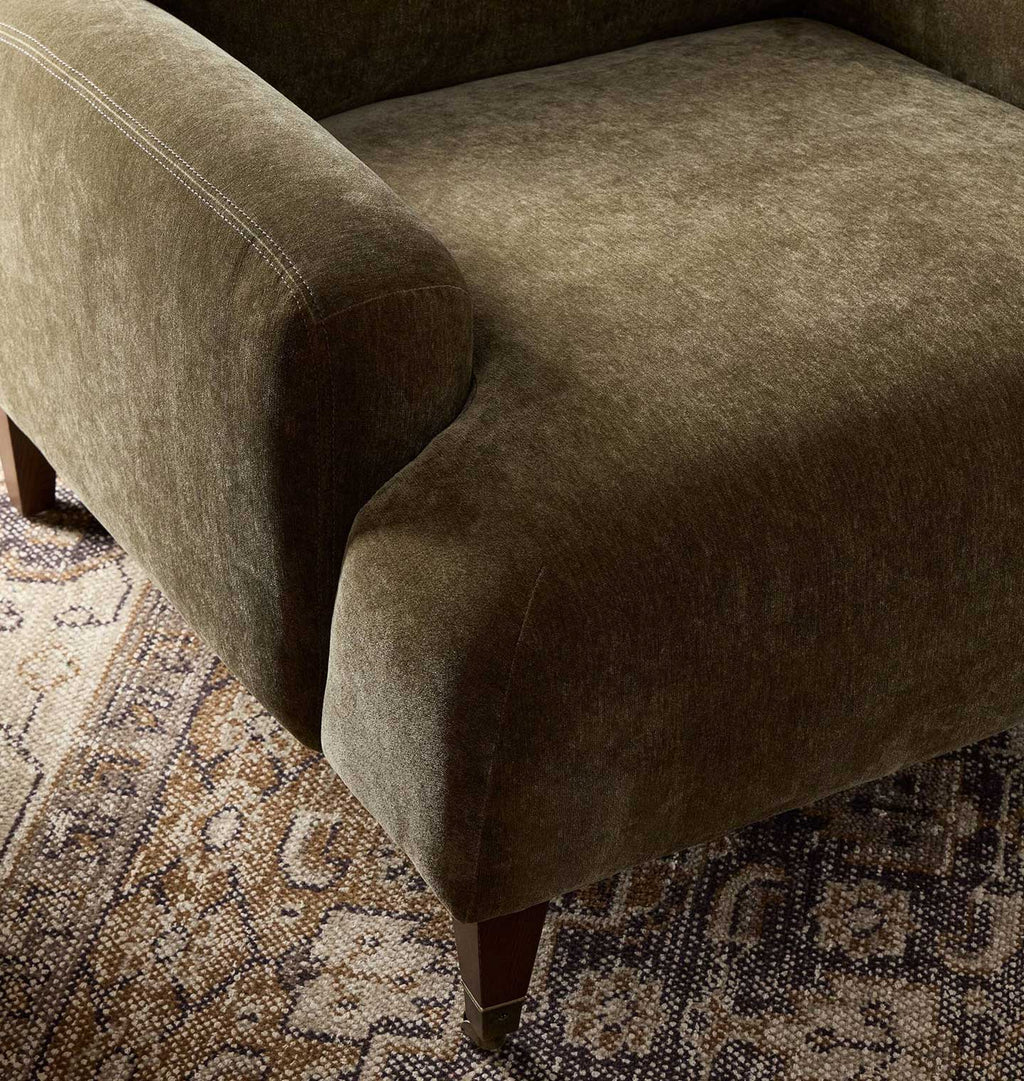 Close-up of an upholstered armchair in a textured taupe-green fabric, showing the seat cushion, armrest, and part of the wooden leg over a patterned rug.