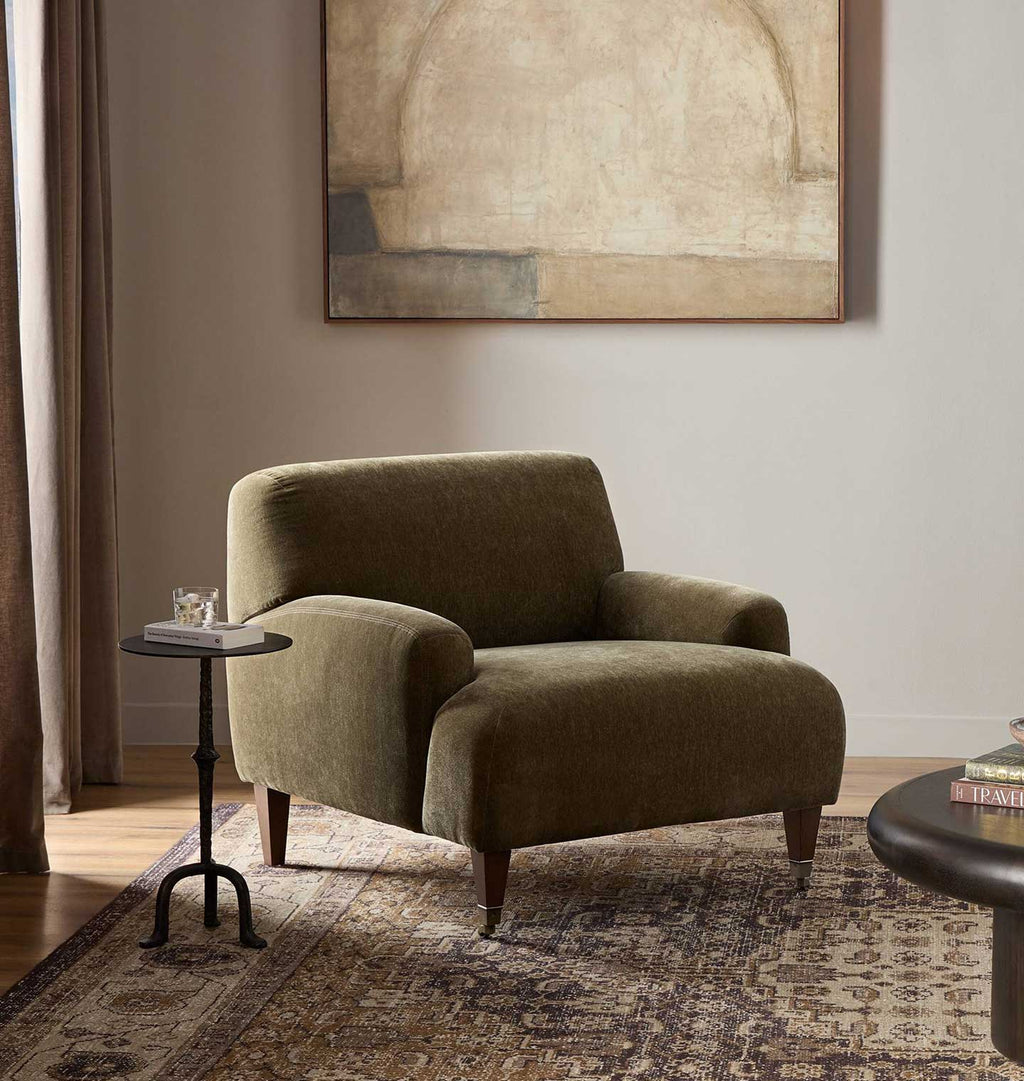 An olive green velvet armchair in a styled living room setting with a patterned rug, a small black side table, and a large abstract painting on the wall.