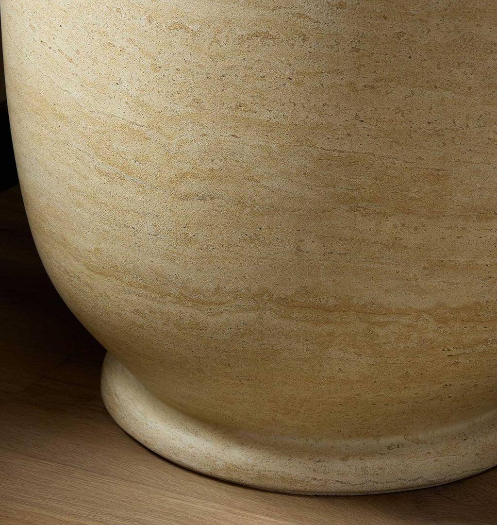 Close-up of a large round stone planter or vase in a warm beige travertine-like finish, resting on a light wood surface.