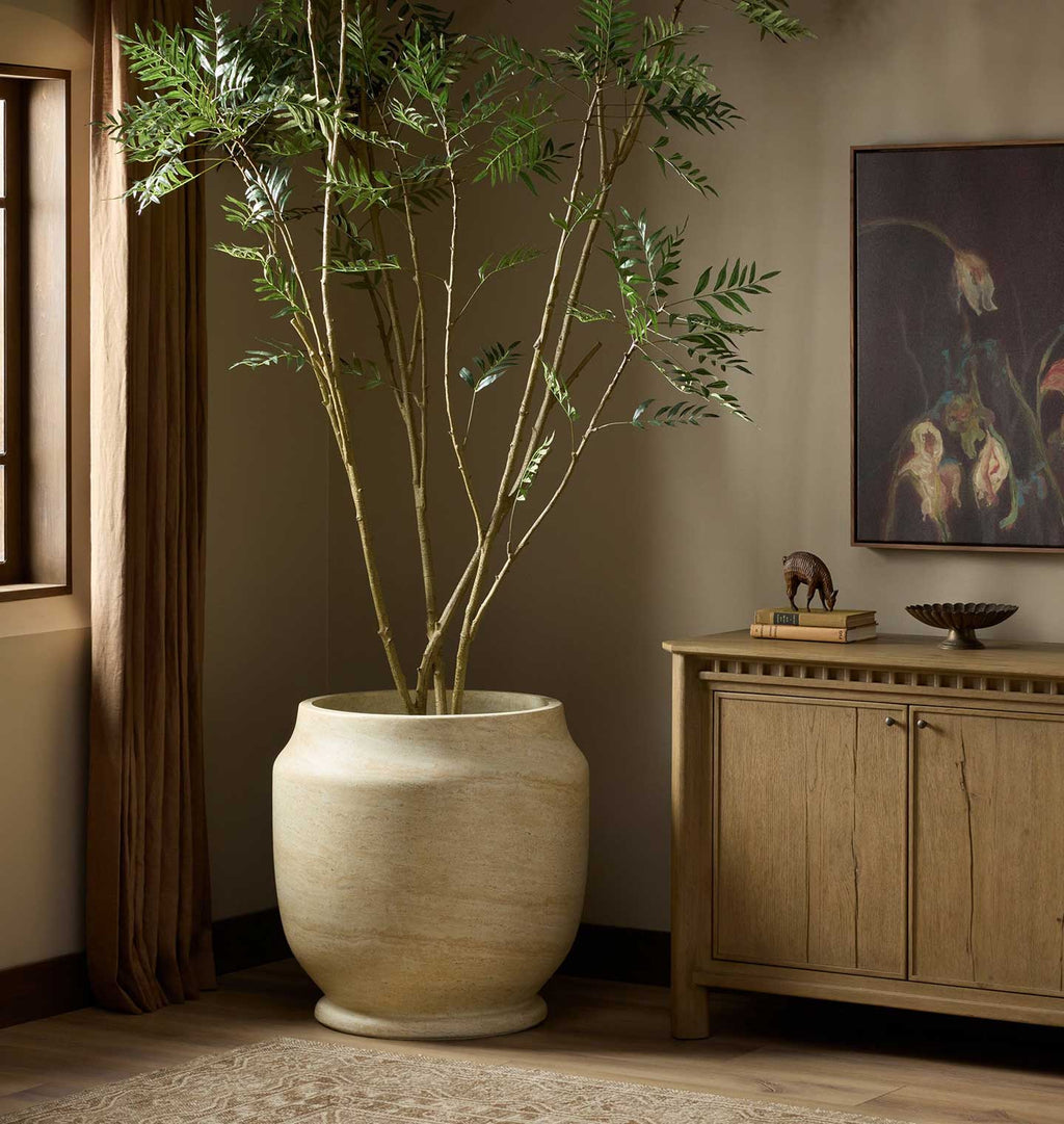 A tall potted artificial bamboo plant with green feathery stems in a large round beige planter, placed in a styled room next to a wooden cabinet and framed artwork.