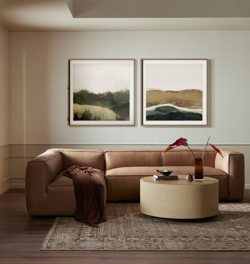 A modern living room featuring a brown leather sectional sofa, a round beige coffee table, and two framed abstract landscape paintings on the wall above the couch.