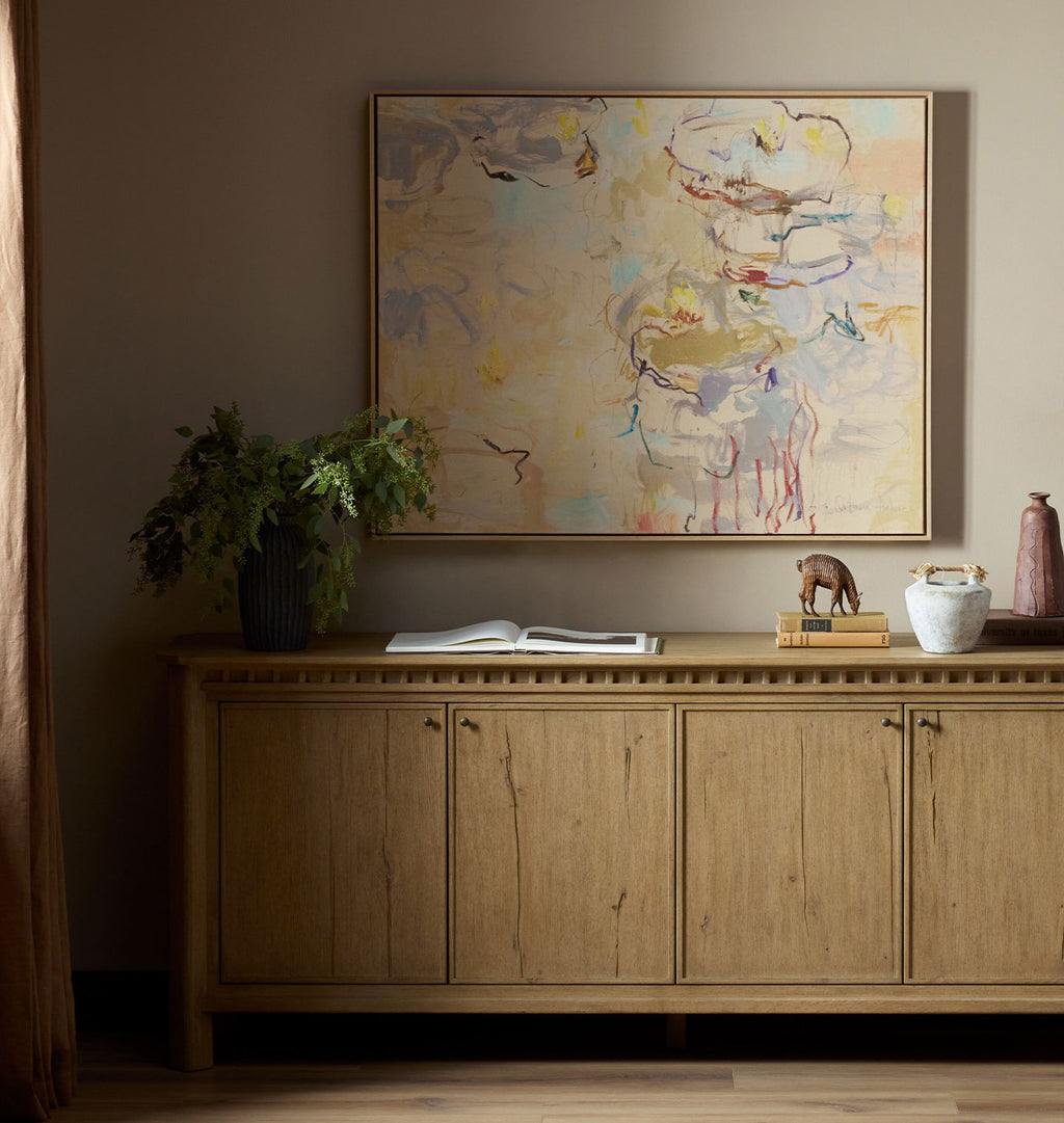 An abstract framed painting hangs on a beige wall above a light wood credenza in a styled room setting with a plant, books, and decorative objects.