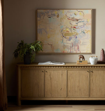 An abstract framed painting hangs on a beige wall above a light wood credenza in a styled room setting with a plant, books, and decorative objects.