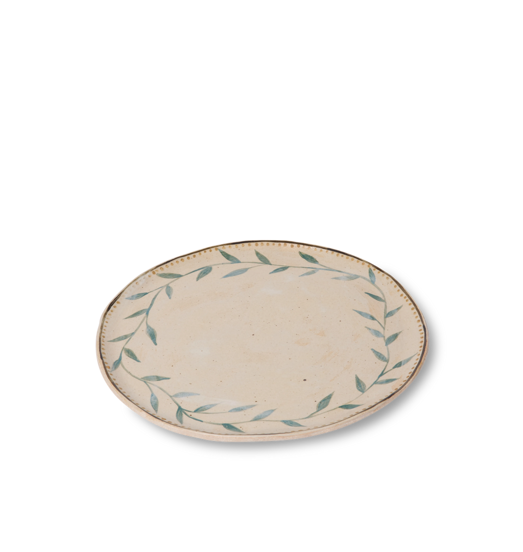 Angled view of a round ceramic plate with an organic rim, decorated with a green leafy vine border and a gold dotted accent line on a white background