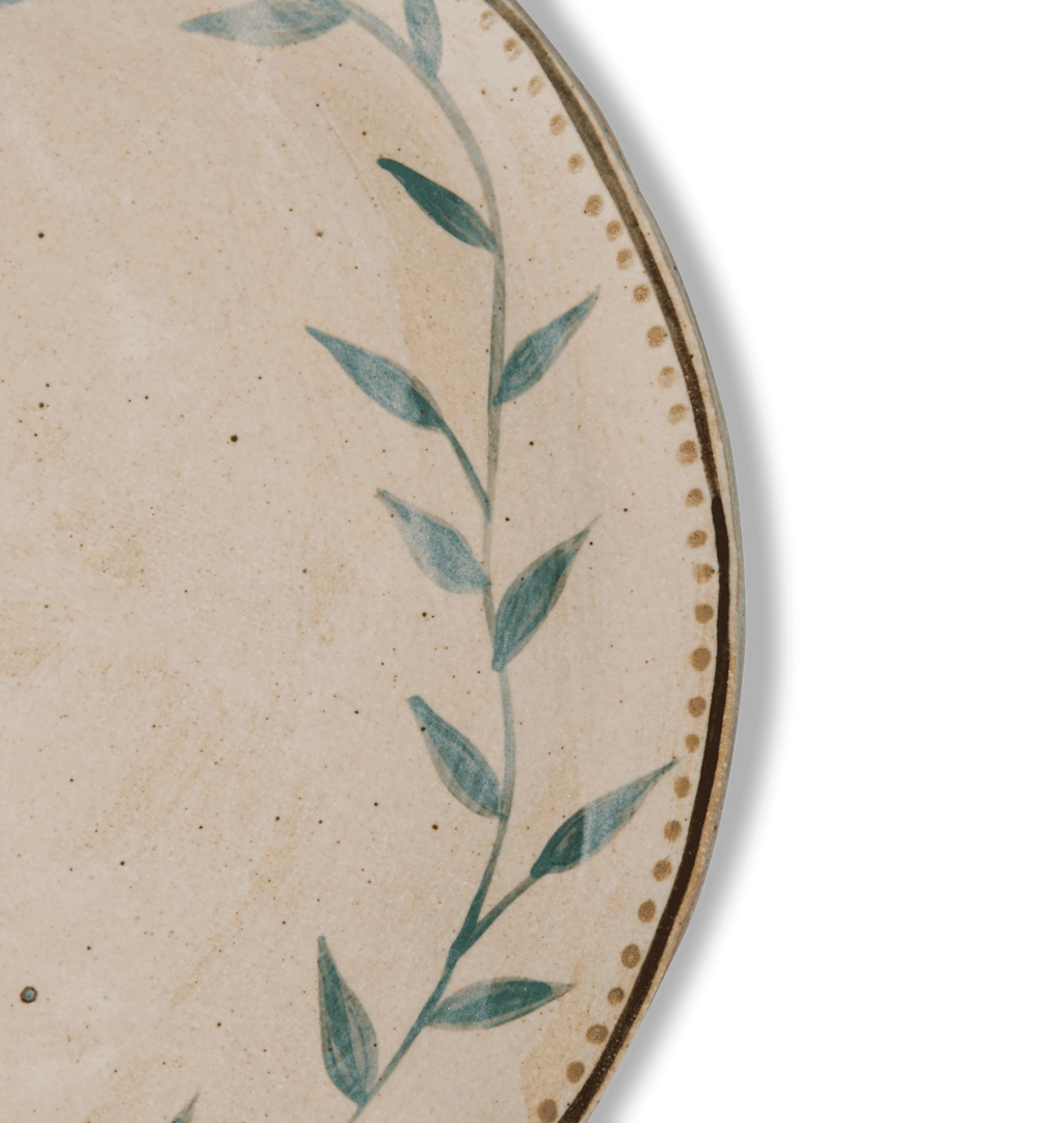 Detailed view of a round ceramic plate with an organic rim, decorated with a green leafy vine border and a gold dotted accent line on a white background