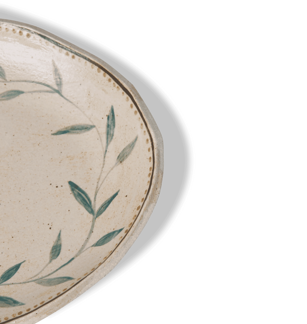 Close up detailed view of an oval ceramic serving platter with an organic rim, decorated with a green leafy vine border and a gold dotted accent line on a white background